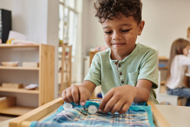 lächelnder afrikanischer amerikanischer Junge, der in der Montessori-Schule mit Tuch und Knöpfen spielt - Foto, Bild
