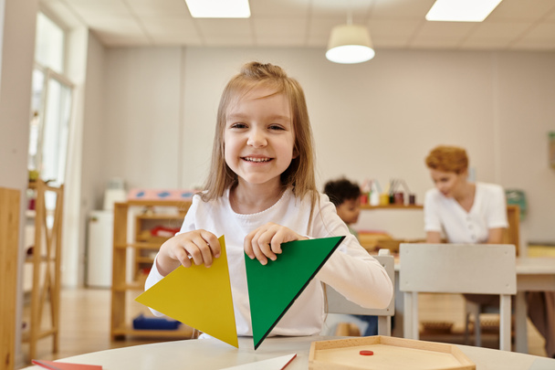 garoto alegre segurando triângulos e olhando para a câmera na aula na escola montessori - Foto, Imagem