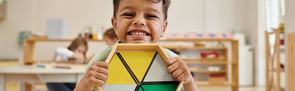 šťastný africký americký chlapec drží dřevěnou hru v rozmazané montessori školy, banner - Fotografie, Obrázek