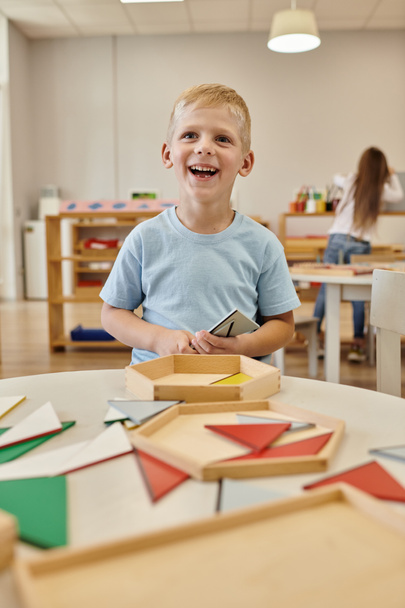 cheerful boy holding wooden triangle and looking away in class in montessori school - Zdjęcie, obraz