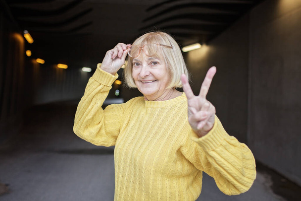 Portrait of woman over 65 with eyeglasses in yellow autumn sweater shows victory gesture. She is cheerful and positive. Beautiful aging and an optimistic outlook on life. - Photo, Image