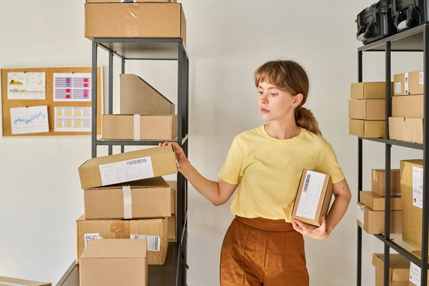 Mujer joven trabajadora de mercado o almacén de pie por rack con paquetes embalados y en busca de caja con el número necesario - Foto, Imagen