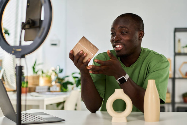 Glücklicher junger Handwerker oder Designer zeigt dem Online-Publikum handgefertigte Vasen, während er im Livestream vor dem Smartphone-Bildschirm sitzt - Foto, Bild