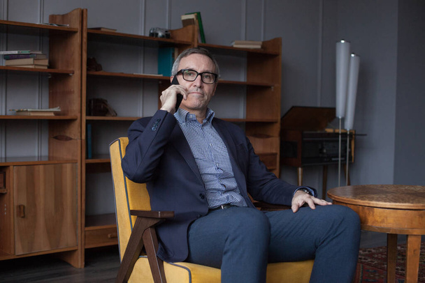 Nervioso hombre de negocios serio en traje azul y gafas hablando en su teléfono móvil en fondo gris estudio de pared - Foto, Imagen