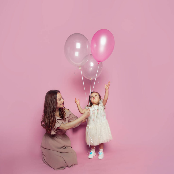 Alegre adorable madre e hija con balón rosa sobre fondo de estudio rosa. Retrato de mamá joven y niña linda bebé - Foto, Imagen