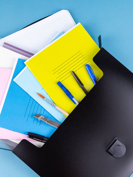 Черный пластиковый портфель папки для документов и школьных предметов на ярко-синий стол рядом с канцелярскими принадлежностями - Фото, изображение