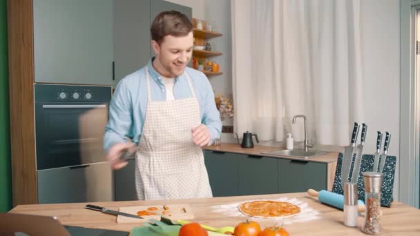 hombre alegre con delantal prepara pizza en la cocina moderna. Macho pone tomates en la masa con la salsa y toma fotos con su teléfono - Metraje, vídeo