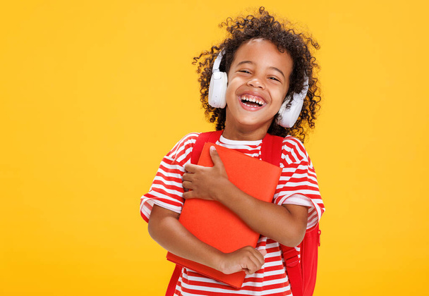 Африканський американський смішний хлопчик з книгою в смугастій футболці регулювання навушників під час прослуховування онлайн-урок після шкільного навчання на жовтому фоні - Фото, зображення