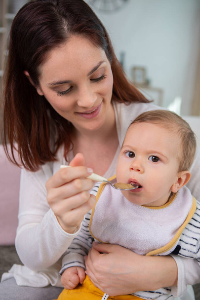 mother feeding baby with spoon indoors - Фото, изображение