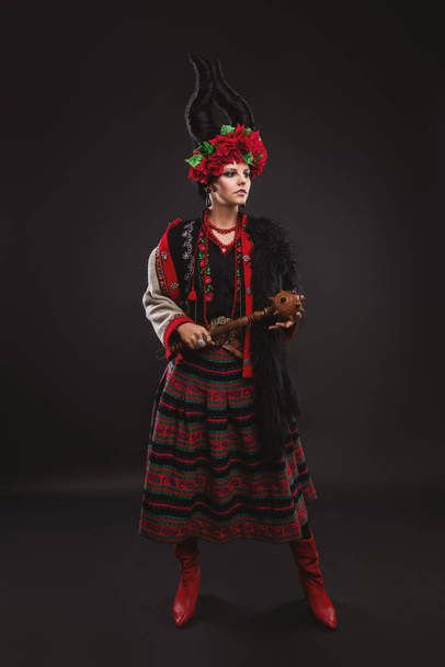 beautiful brunette woman in a traditional Ukrainian costume, with a floral wreath and horns, is standing on the black studio background - Photo, Image
