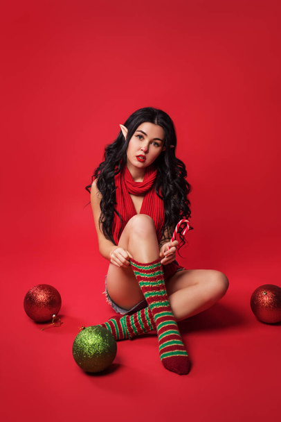 sexy young woman in elf costume and with elven ears is standing on the red studio background with a Christmas balls, pin-up style, new year sale - Fotó, kép