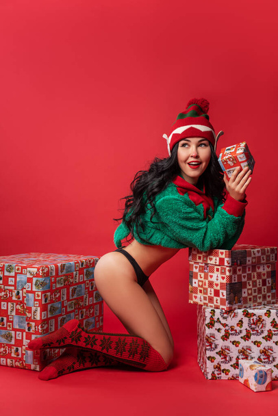 sexy young woman in elf costume and with elven ears is standing on the red studio background with a Christmas gift, pin-up style, new year sale - Fotoğraf, Görsel