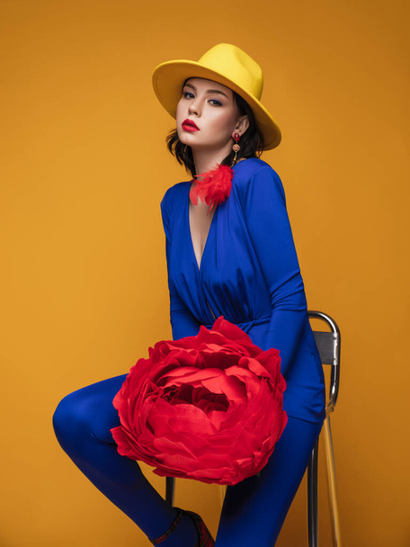 portrait studio de mode d'une belle femme en costume bleu, chapeau jaune, plume rouge et avec du papier géant rose sur le fond jaune - Photo, image