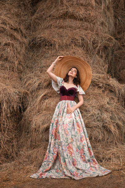 beautiful young brunette woman in a long retro dress and a big straw hat standing near the hayloft - Zdjęcie, obraz