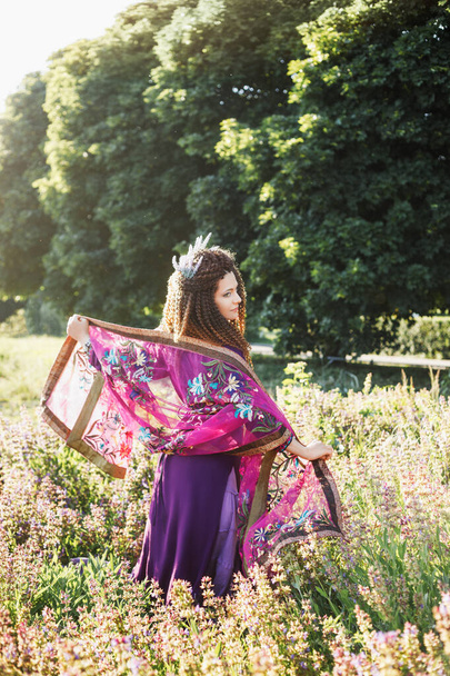 Mor etnik elbise içinde kıvırcık saçlı ve mor makyajlı çekici esmer kadın ve lavanta çiçekli taç dans ediyor ve lavanta çiçekleri içinde gülümsüyor. - Fotoğraf, Görsel