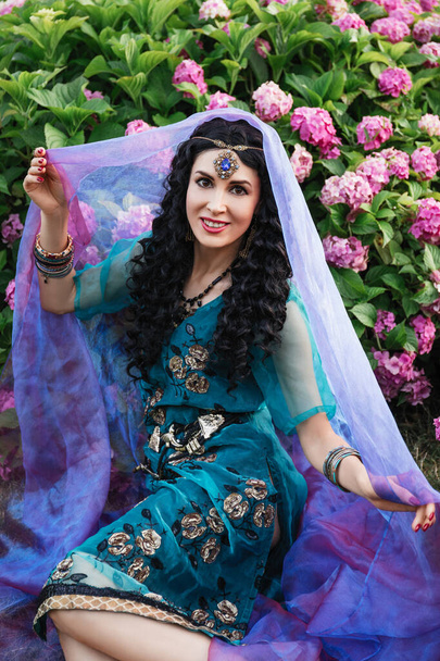 piękna brunetka księżniczka kobieta z kręconymi włosami i diadem na głowie, w niebieskiej indyjskiej sukience sari, siedzi w ogrodzie w kwiaty z przezroczystą tkaniną w jej rękach i uśmiechnięty - Zdjęcie, obraz