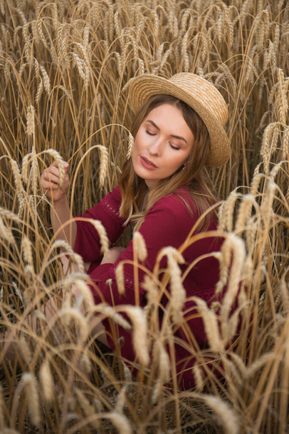 Een jonge vrouw in het tarweveld met een spikelet in haar hand. Dame met strohoed en boho stijl rode jurk. - Foto, afbeelding