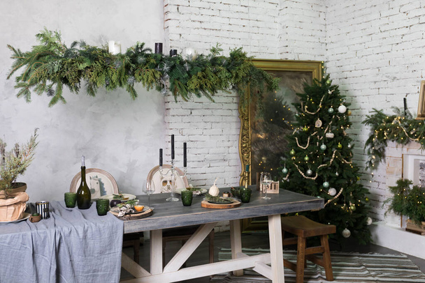 Wohnzimmer Esszimmer im Loftstil mit Ziegelwänden, einem alten Spiegel im Rahmen, einem Weihnachtsbaum, einem großen Esstisch mit Holzbänken, einer blauen Tischdecke, einem gestreiften Teppich, Tellern,   - Foto, Bild