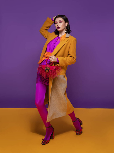красивая брюнетка с цветовым макияжем в фиолетовом платье и оранжевом поясе, в желтом пальто и красной обуви, с красной сумкой и серёжкой из желтых перьев стоит на фиолетовой и желтой студии - Фото, изображение