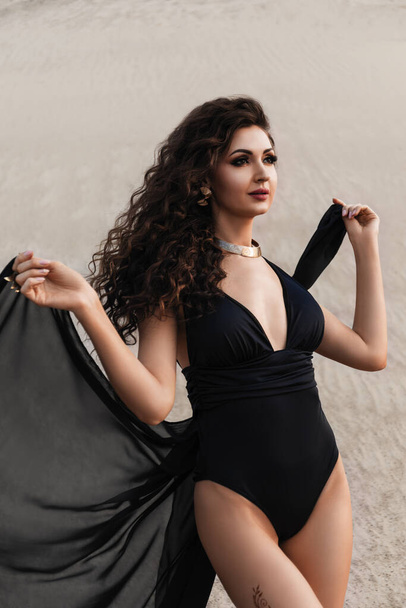beautiful sexy young brunette woman with curly hair and with evening makeup, in black swimsuit and with transparent fabric, mehendi, golden accessories, in desert sands on the beach - Photo, Image