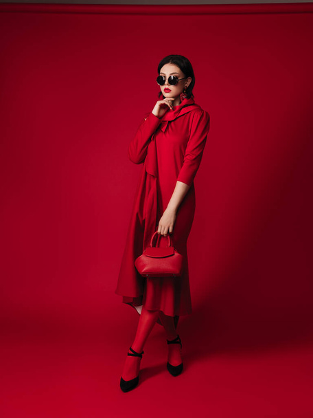 total red look style fashion studio portrait of a gorgeous young brunette woman, coat, bag, glasses, head scarf, makeup, brooch with feathers, red lips, high heels - Photo, Image