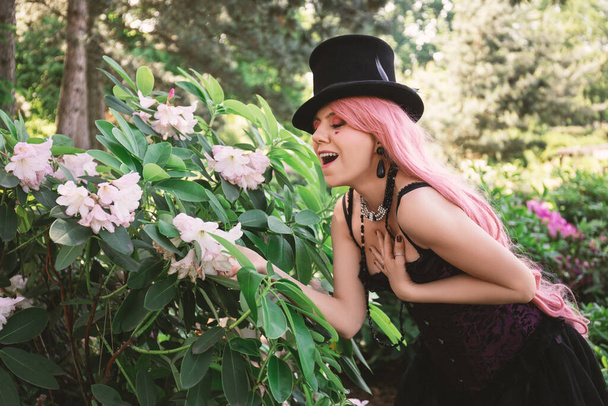 όμορφη γυναίκα με ροζ μαλλιά, σε ψηλό καπέλο και σε βραδινό μαύρο φόρεμα με κορσέ, κάθεται στον κήπο με λουλούδια αζαλέας - Φωτογραφία, εικόνα