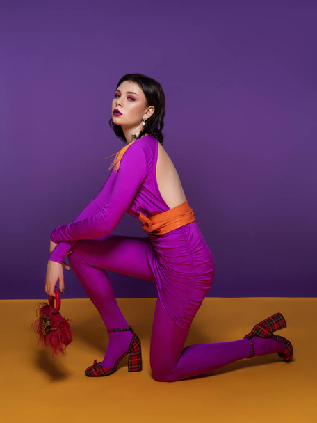hermosa mujer morena con maquillaje de color brillante en un vestido púrpura y un cinturón naranja, en zapatos de tacón alto de cuadros rojos, con una bolsa roja y un pendiente de plumas amarillas  - Foto, Imagen