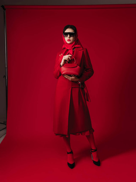total red look style fashion studio portrait of a gorgeous young brunette woman, coat, bag, glasses, head scarf, makeup, brooch with feathers, red lips, high heels - Foto, afbeelding