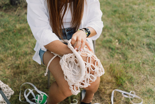 女性はテントの近くの屋外でマクラメ技術を使用してバッグを編みます. アウトドア趣味。 マクラム・ウィービング. - 写真・画像