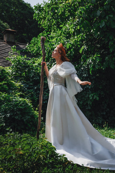 belle femme aux cheveux roux dans un costume médiéval blanc, dans une couronne et avec un bâton se dresse dans une forêt verte, princesse fantaisie - Photo, image