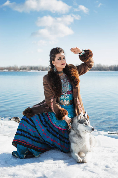 bela mulher morena com cão husky cinza está sentado na neve, penteado de trança, traje de estilo étnico boho azul e rosa, casaco de pele marrom, maquiagem à noite, acessórios de prata, inverno, lago, céu - Foto, Imagem