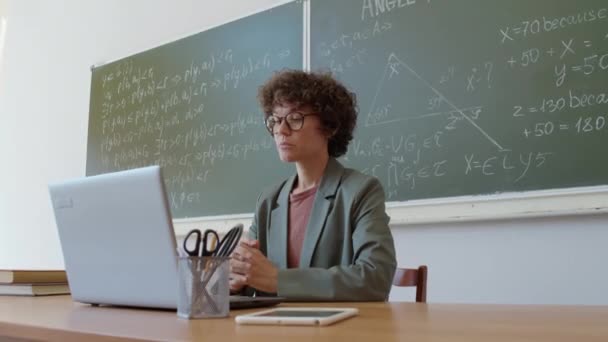 Средний портрет преподавателя колледжа, сидящего за партой перед ноутбуком и объясняющего предмет студентам во время онлайн-урока - Кадры, видео