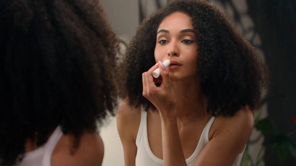 Beautiful African American woman looking in mirror reflection morning beauty routine preparation ethnic girl in bathroom applying moisturizing lip balm lipstick smooth lips organic natural cosmetic - Photo, Image
