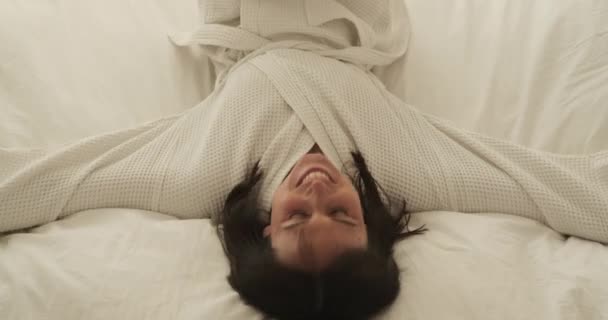 Happy woman is seen playfully falling onto the bed with a joyful smile. Her radiant expression reflects her sense of delight and contentment. Soft lighting adds to the cheerful ambiance. - Footage, Video