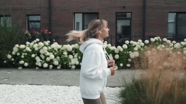 Вид збоку повільний рух сучасної молодої жінки, що слухає музику в бездротових навушниках під час бігу вранці - Кадри, відео