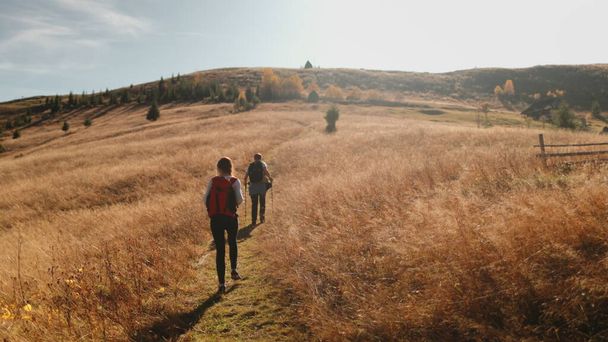 Tourists walking autumn mountain trekking trail on yellow grass meadow. Man and woman couple backpack hiking together in sunset summer highlands. Travel, tourism, holiday, active vacation. Slow motion - Photo, Image