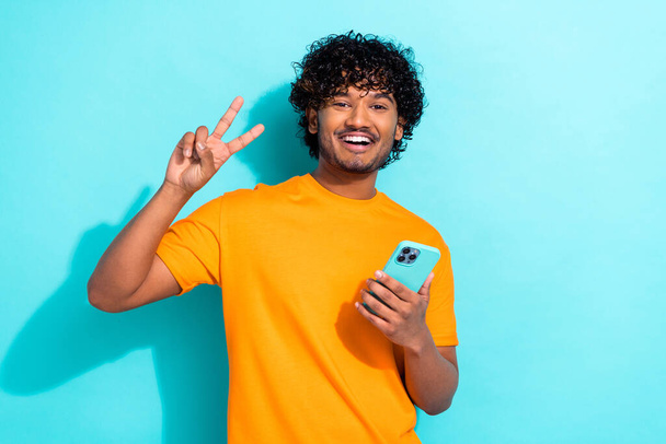 Photo of funky positive handsome man toothy smile hold smart phone hand fingers demonstrate v-sign isolated on teal color background. - Foto, afbeelding