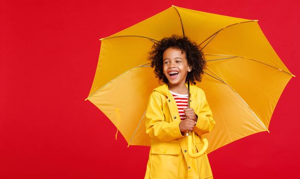 Yağmurluk giymiş neşeli Afro-Amerikalı çocuk kırmızı arka planda, yağmurlu bir günde sırtına sarı şemsiye takıyor ve gülüyor. - Fotoğraf, Görsel