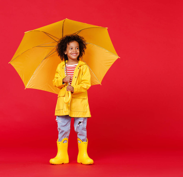 Engraçado menino americano africano em capa de chuva e botas com guarda-chuva de pé enquanto caminhava contra o pano de fundo vermelho no dia chuvoso - Foto, Imagem