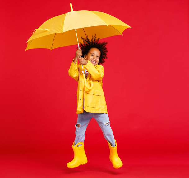 Yağmurlu bir günde kırmızı zeminde yürürken şemsiyeli ve botlu komik Afro-Amerikalı çocuk zıplıyor. - Fotoğraf, Görsel