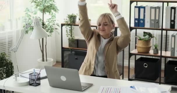 Office worker joyfully dancing while sitting at desk in the office. With a lively and carefree demeanor, she adds a touch of fun to her workday. Despite her dance, she remains focused on her tasks. - Footage, Video