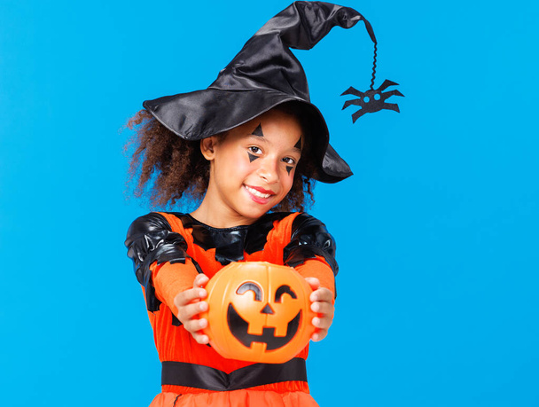 Heureuse fille afro-américaine joyeuse avec des cheveux bouclés en costume de citrouille célèbre Halloween et rit sur fond bleu vif - Photo, image