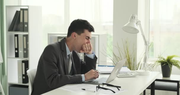 El cansado oficinista caucásico es capturado bostezando mientras está sentado en su escritorio. Su expresión facial refleja fatiga, y el bostezo indica la necesidad de descanso y rejuvenecimiento. - Metraje, vídeo