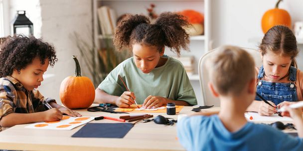 Onnellinen hymyilevä monikansallinen ryhmä lapsia, jotka tekevät Halloween-sisustuksia yhdessä, lapset maalaavat kurpitsoja ja tekevät paperisia pistokkaita. - Valokuva, kuva
