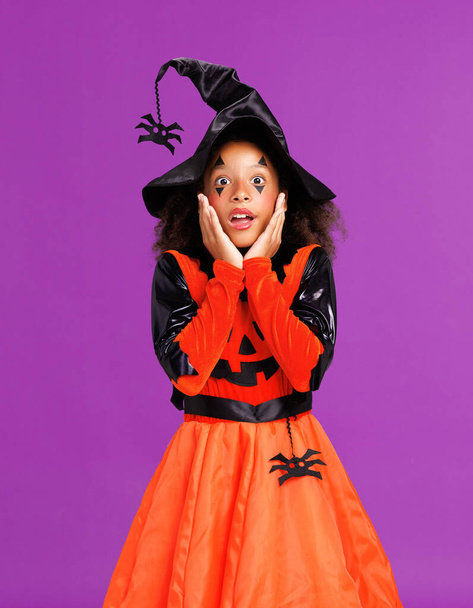 Shocked cheerful african american girl with curly hair  in costume pumpkin touches cheeks in surprise  while  celebrates Halloween on  bright purple background - Foto, afbeelding
