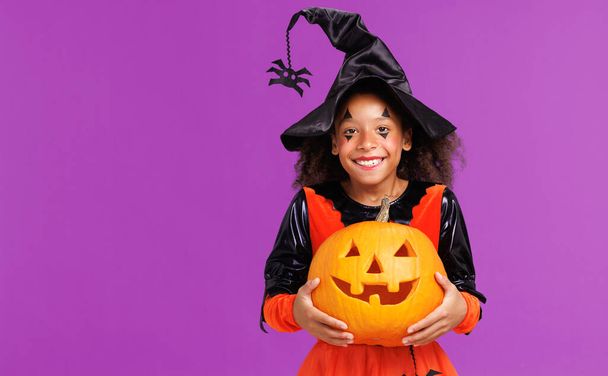 カボチャの衣装のカーリーヘアを持つ幸せな陽気なアフリカ系アメリカ人の少女はハロウィーンを祝い,明るい紫色の背景に笑います - 写真・画像