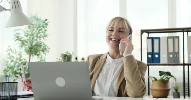 Hymyilevä naisyrittäjä osallistuu ystävälliseen puhelinkeskusteluun ystäviensä kanssa työpaikallaan. Lämmin ja iloinen käytös, hän näyttää nauttivan keskustelusta. - Materiaali, video