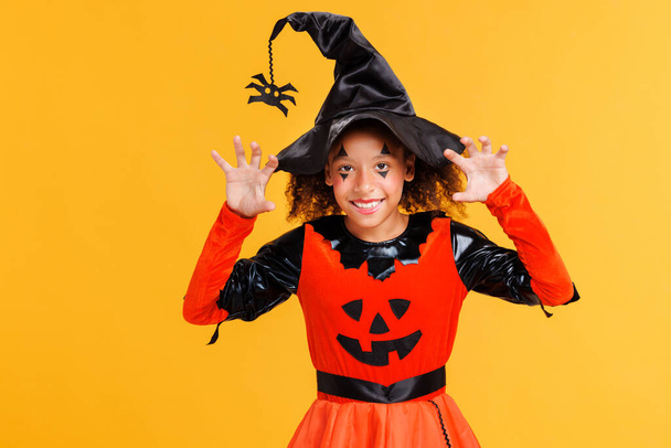 Heureuse fille afro-américaine joyeuse avec des cheveux bouclés en costume de citrouille gestes effrayants et célèbre Halloween et rit sur fond jaune - Photo, image