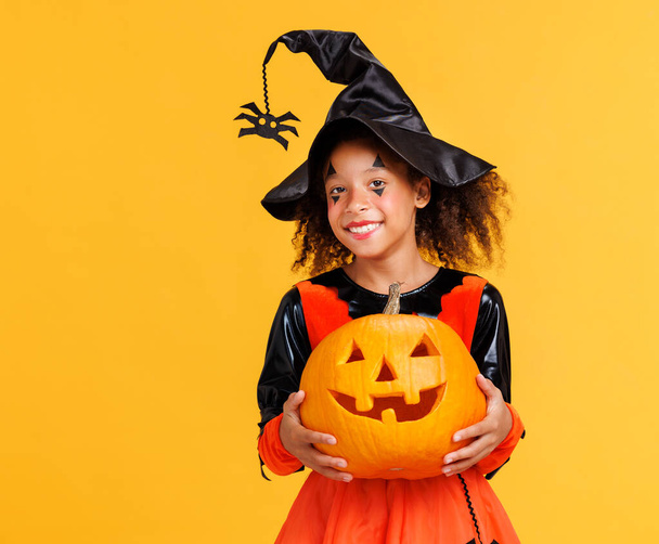 Happy cheerful african american girl with curly hair in  pumpkin costume with jack-o-lantern celebrates Halloween and laughs on yellow background - Photo, Image