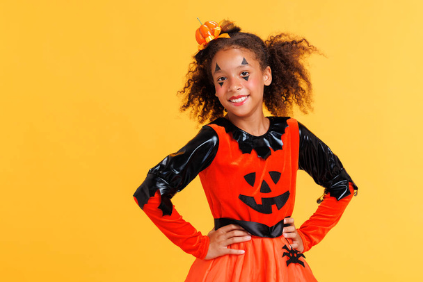 Heureuse fille afro-américaine joyeuse avec des cheveux bouclés en costume de citrouille célèbre Halloween et rit sur fond jaune - Photo, image